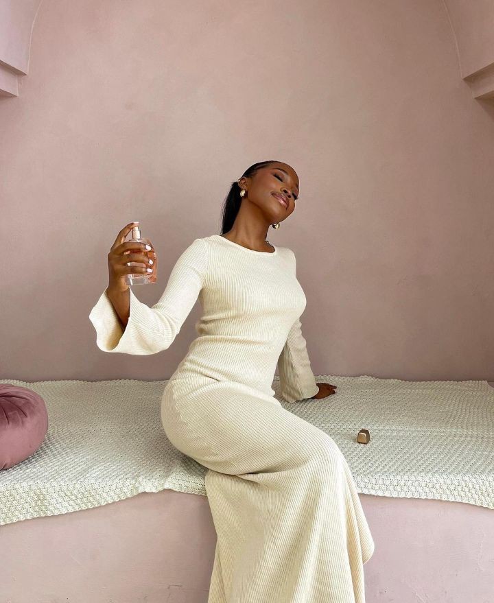 Amy Okoli is wearing cream coloured, long sleeved dress, holding a perfume bottle in one hand while reclining gracefully.